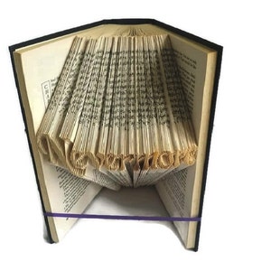 May include: A book with pages folded into the shape of the word "LOVE" in a 3D design. The book is closed and has a black cover. A purple rubber band is wrapped around the book.