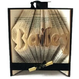 May include: A graduation themed book art with the name "Peter" folded into the pages. The book is framed with a black border and a black ribbon with a gold tassel.