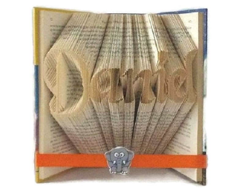 May include: A book with the name "Darla" folded into the pages. The book is open and the pages are fanned out to create a 3D effect. A small elephant figurine is attached to the front of the book.
