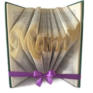 Könnte beinhalten: Ein Buch, dessen Seiten zu dem Wort "Love" gefaltet sind, mit einem violetten Band darum.