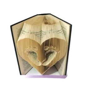 May include: A book with pages folded to create a heart shape, with baby footprints cut out within the heart. The book has a black spine and the pages are a light tan color. The book is open, and the heart is visible.