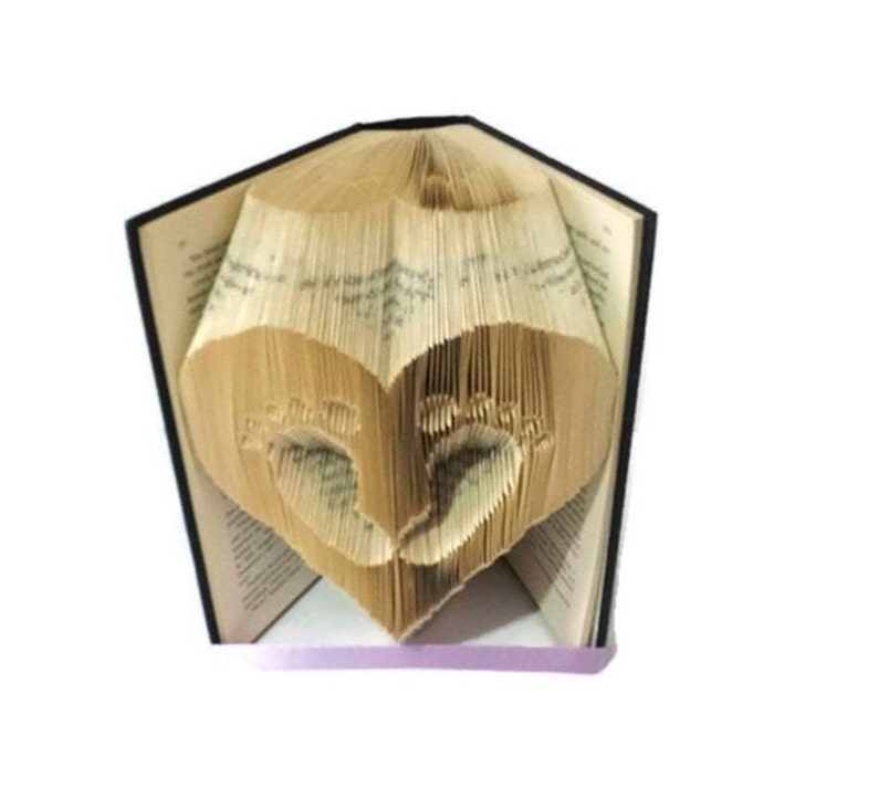 May include: A book with pages folded to create a heart shape, with baby footprints cut out within the heart. The book has a black spine and the pages are a light tan color. The book is open, and the heart is visible.