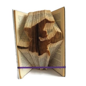 Op de afbeelding: Een boekensculptuur van een kat, gemaakt door de pagina's van een boek te vouwen. Het boek is open en de kat staat op zijn achterpoten. Het boek wordt bij elkaar gehouden met een paarse elastiek.