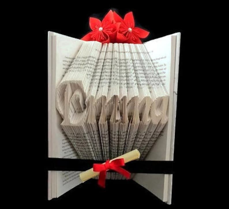 May include: A book art sculpture with the word "Drama" folded into the pages. The book is decorated with a red flower on top and a red ribbon and scroll at the bottom.