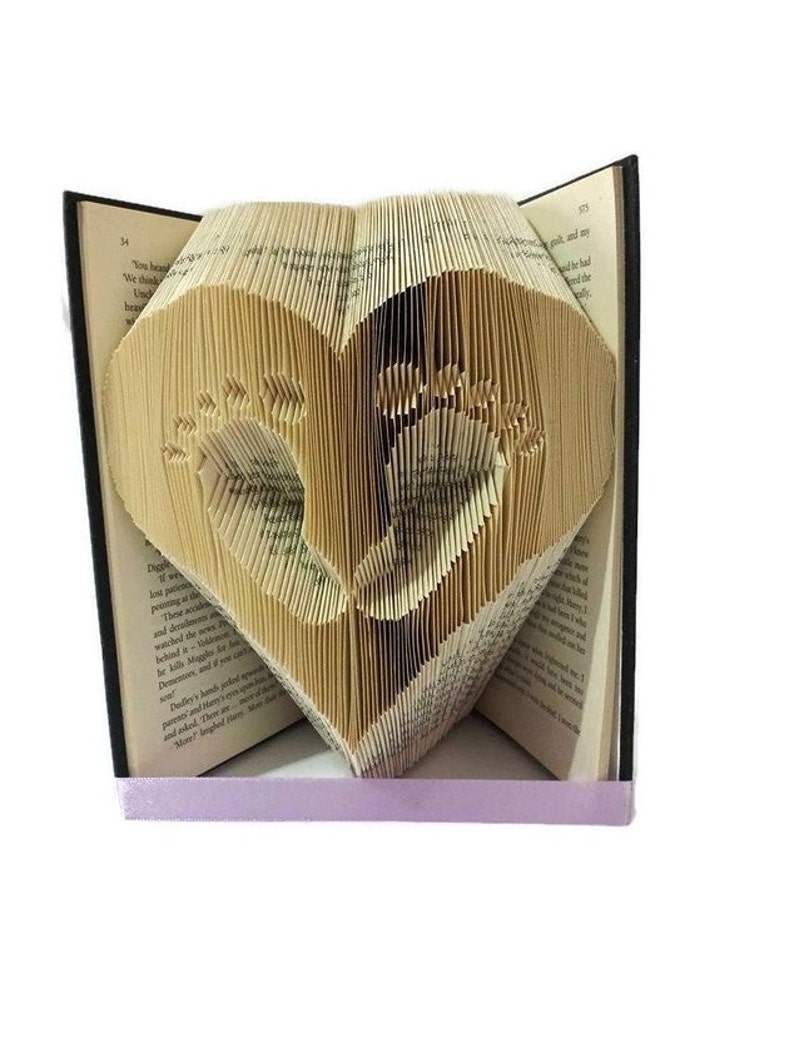 May include: A heart-shaped book sculpture with baby footprints cut into the pages. The book is open, with the heart design centered. A lavender ribbon is at the base. The pages are aged, and the book has a black spine.