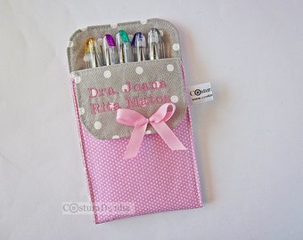 May include: A gray and pink fabric pencil case with white polka dots. The case has a pink ribbon and is personalized with the text "Dra. Joana Rita Matos".