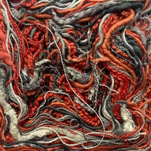 May include: Close-up of a textured textile art piece. The artwork features a complex arrangement of various yarns in shades of red, orange, gray, and white. The yarns are intertwined, creating a dense and tactile surface, with visible textures and patterns.