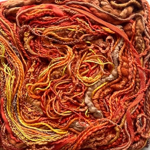 May include: Close-up of a vibrant ball of yarn in shades of orange, yellow, and brown. The yarn has a variety of textures, including thick and thin strands, braids, and ribbons. The colors blend together to create a warm, inviting look, perfect for knitting or crochet projects.