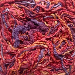 May include: Close-up of a tangled mass of colorful yarn. The yarn features a variety of textures and shades, including deep reds, oranges, purples, and pinks. The different strands are intertwined, creating a rich and complex visual.