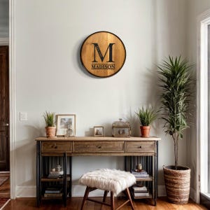 May include: A wooden round sign with a black letter "M" and the text "EST. 2023 MADISON" in black lettering. The sign is hanging on a white wall above a wooden desk with drawers and shelves. A potted plant is on the left side of the desk and a larger potted plant is on the right side of the desk.