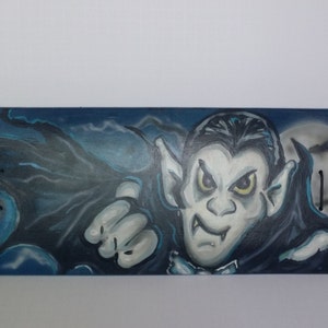 Op de afbeelding: Een blauw skateboarddeck met een handgeschilderd ontwerp van een vampier met tanden en vleermuisvleugels. De achtergrond is donkerblauw met witte wolken en vleermuizen.