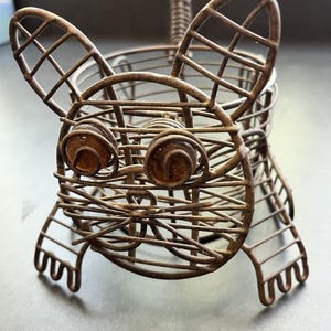 May include: A whimsical, wire-frame sculpture of a dog. The sculpture is made of rusty brown metal and features large ears, round eyes, and a basket-like body. The dog is standing on four legs with a looped design.