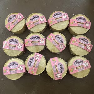 May include: A collection of twelve round, gold-colored mystery dumpling toys. Each toy is wrapped with a pink band featuring the text "Mystery Dumpling" and "Collect All 12 Colors". The toys are designed for children aged 3 and up.