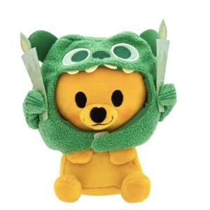 Könnte beinhalten: Ein gelber Teddybär mit einer grünen Monsterkapuze mit weißen Augen und Zähnen. Der Bär hält zwei weiße Papierkerzen mit gelben Flammen. Das Spielzeug hat einen runden Körper und ist vor einem weißen Hintergrund platziert.