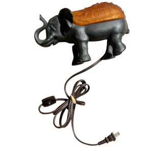 May include: A gray elephant-shaped lamp with a brown saddle. The lamp has a brown electrical cord with a switch and plug. The elephant has large ears and a trunk. The saddle has a textured design.