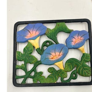 May include: A cast iron wall decor piece featuring blue morning glory flowers with yellow centers and green leaves. The flowers are set against a white background within a black rectangular frame. A decorative garden accent.