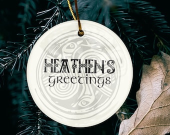 May include: A round, off-white Christmas ornament with the text "HEATHEÑS greetings" in a Celtic-inspired font. The ornament is suspended by a gold cord against a backdrop of dark green pine needles and a dried leaf.