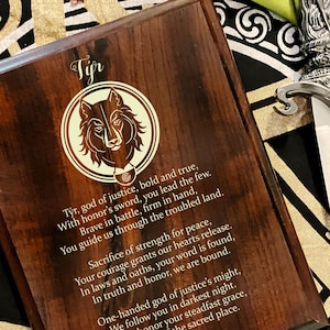 May include: A wooden plaque with a wolf head design and an inscription about Tyr, the Norse god of justice, honor, and strength. The inscription reads: "Tyr, god of justice, bold and true, With honor's sword, you lead the few. Brave in battle, firm in hand, You guide us through the troubled land. Sacrifice of strength for peace, Your courage grants our hearts release. In laws and oaths, your word is found, In truth and honor, we are bound. One-handed god of justice's might, We follow you in darkest night. Týr, we honor your steadfast grace, Protector of the sacred place. Hail Tyr, so it is."