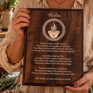 May include: A rectangular dark wood plaque featuring an engraved Hestia design with a flame in a bowl, a Greek key border, and a poem titled Hestia. The text reads: Hestia, Keeper of the Hearth, First and last among the sacred ones, Your flame burns steady, a beacon of home, A warmth that welcomes and binds us together. Guardian of sacred fire, You who dwell in every kindled hearth, Bless those who gather in peace and love, And grant solace to all who seek refuge. Teach us the virtue of stillness, The quiet strength of devotion and care, That in our homes, your flame may never fade. Hail Hestia, in your warmth we dwell.