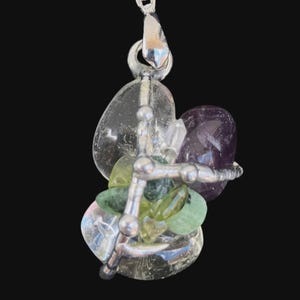May include: A silver pendant necklace featuring a cluster of polished gemstones. The pendant includes clear, green, and purple stones, held together by a silver metal structure. The necklace hangs from a dark gray cord.
