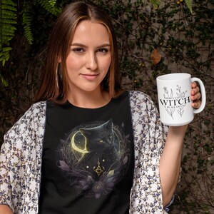May include: A woman wearing a black t-shirt with a black cat and moon graphic holds a white mug with the text "There's a little witch in all of us".