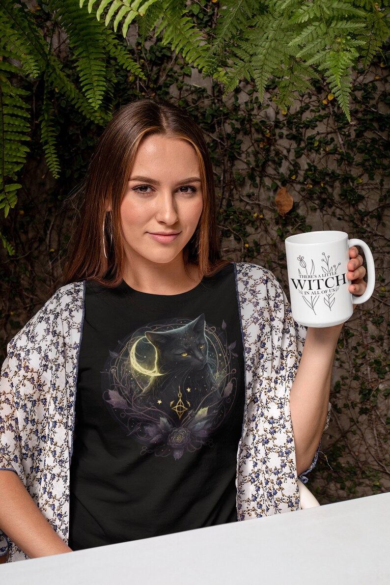 May include: A woman wearing a black t-shirt with a black cat and moon graphic holds a white mug with the text "There's a little witch in all of us".