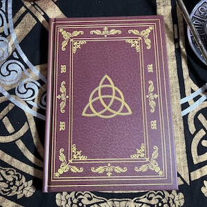 Peut inclure: Un livre de couleur bordeaux avec une bordure dorée décorative et un symbole de nœud celtique doré. La couverture a un aspect texturé. Le livre est rectangulaire et semble fermé. Le fond est un tissu à motifs.