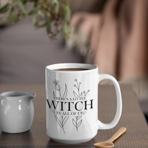 May include: White ceramic mug with a black outline of flowers and the text "There's a little witch in all of us".