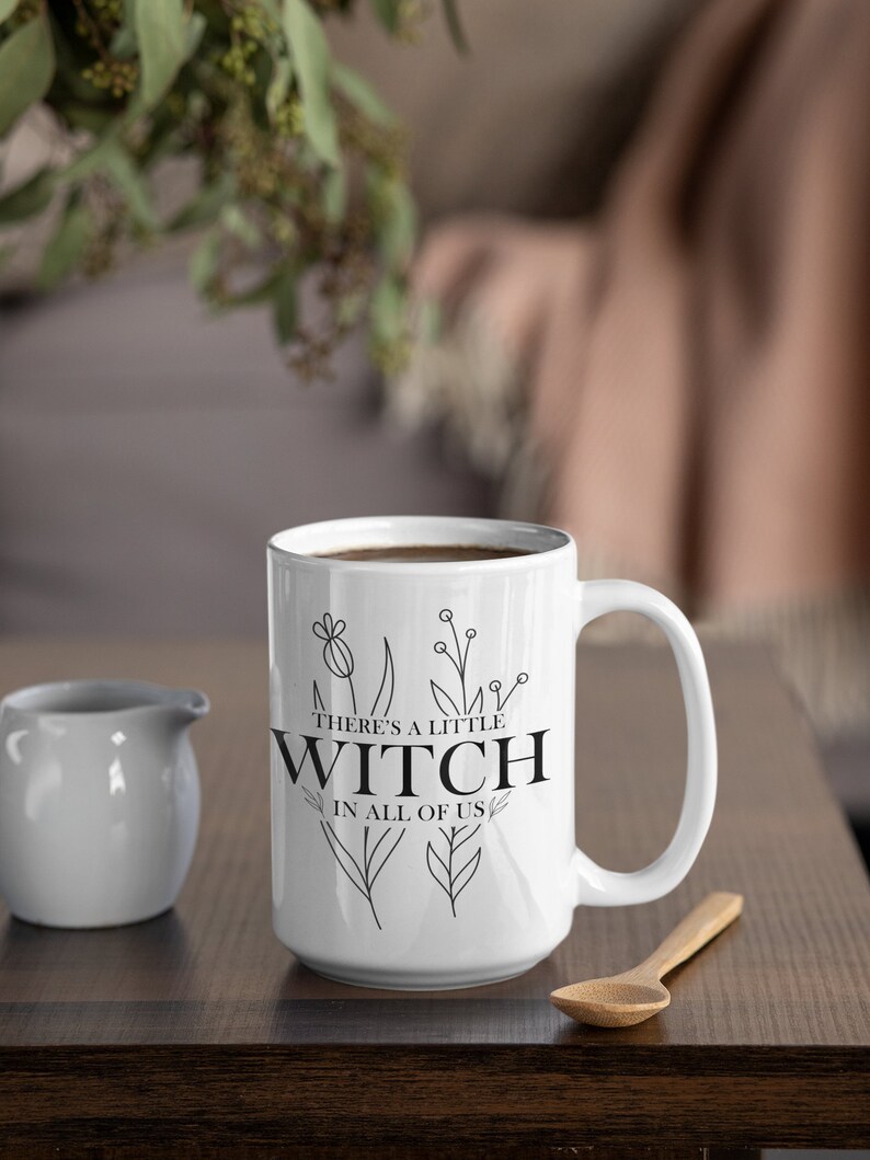 May include: White ceramic mug with a black outline of flowers and the text "There's a little witch in all of us".