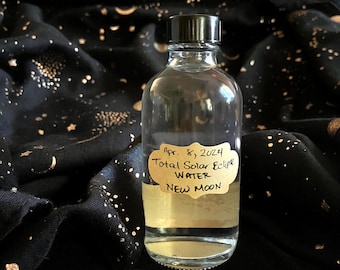 May include: A clear glass bottle with a black cap, containing a liquid. The bottle has a label that reads "Apr. 8, 2024 Total Solar Eclipse Water New Moon".