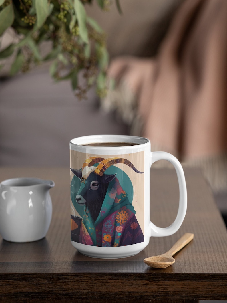 May include: A white ceramic mug featuring a whimsical illustration of a goat wearing a colorful robe. The goat has large golden horns and is set against a teal circle. A small white creamer and wooden spoon are on the table.
