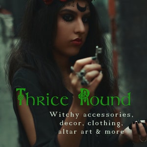 May include: A woman wearing a black outfit and a crown of red roses holds a silver key. The text "Thrice Round" is written in green with a Celtic knot design. The text below reads "Witchy accessories, decor, clothing, altar art & more".