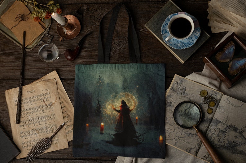 May include: A tote bag with a dark blue background featuring a woman in a red cloak standing in a snowy forest. She is holding a glowing orb in her hand and there are candles on the ground around her.