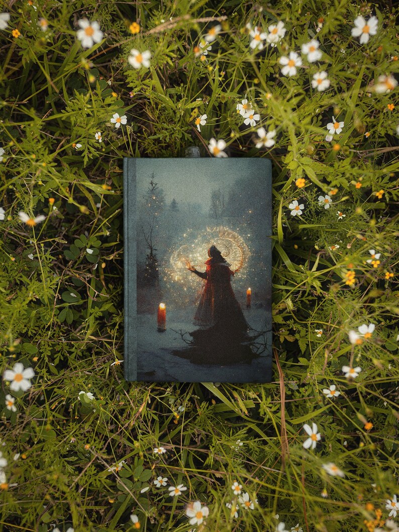 May include: A hardcover book with a dark blue cover featuring a woman in a red cloak standing in a forest with glowing lights. The book is surrounded by white flowers.