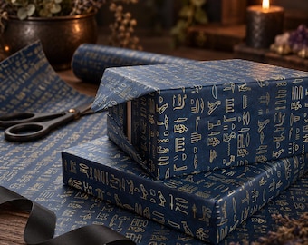 Puede incluir: Una caja de regalo rectangular envuelta en papel azul oscuro con un patrón repetitivo de color dorado de jeroglíficos egipcios antiguos. La caja descansa sobre una hoja de papel de regalo a juego junto a unas tijeras negras y una cinta negra.