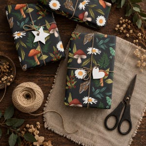 Peut inclure: Trois cadeaux rectangulaires emball&eacute;s dans du papier noir avec un motif de champignons rouges, de marguerites blanches, de foug&egrave;res vertes et de papillons de nuit. Les paquets sont li&eacute;s avec de la ficelle brune et orn&eacute;s d'&eacute;tiquettes en papier blanc.