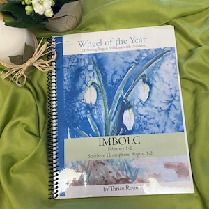 Può includere: Un libro rilegato a spirale intitolato "Wheel of the Year: Exploring Pagan holidays with children." La copertina presenta un dipinto ad acquerello di fiori bianchi di bucaneve su uno sfondo blu. Il testo "IMBOLC" è stampato in grandi lettere sotto i fiori, con le date "February 1-2" e "Southern Hemisphere: August 1-2" stampate sotto. Il libro è di "Thrice Round."