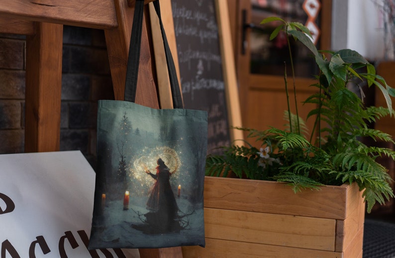 May include: A tote bag with a dark blue background featuring a woman in a red dress holding a glowing orb in a snowy forest setting. The bag has black straps.