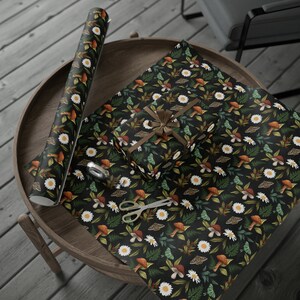 Peut inclure: Un rouleau de papier cadeau et une bo&icirc;te cadeau emball&eacute;e avec le m&ecirc;me papier, pr&eacute;sentant un fond sombre avec un motif r&eacute;p&eacute;titif de marguerites blanches, champignons bruns, foug&egrave;res vertes et papillons de nuit. Des ciseaux argent&eacute;s.
