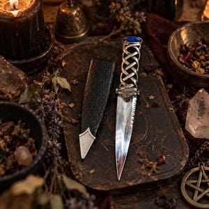 Peut inclure: Un poignard athamé argenté doté d'un manche en métal tressé surmonté d'une pierre ovale bleue, accompagné d'un fourreau noir texturé avec une pointe en métal argenté, posé sur une surface rectangulaire sombre gravée de symboles lunaires.