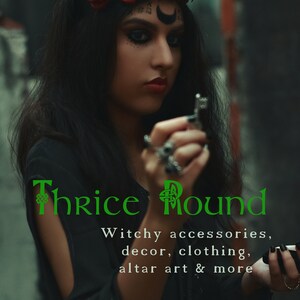 May include: A woman wearing a black outfit with a crown of red roses and candles. The text "Thrice Round" is written in green with a Celtic knot design. The text below reads "Witchy accessories, decor, clothing, altar art & more".