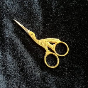 May include: Gold-colored embroidery scissors in the shape of a bird, with a detailed design on the handle and sharp blades. The scissors are set against a dark background.