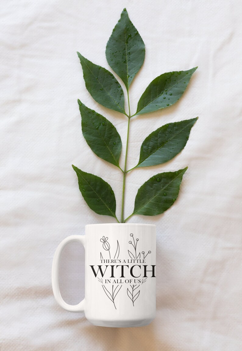 May include: White ceramic mug with a black design that reads "There's a little witch in all of us". The design includes a few stylized flowers. The mug is sitting on a white surface with a green leafy branch behind it.
