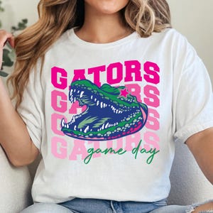 May include: White t-shirt featuring a green and blue alligator graphic with the word "GATORS" in pink and the words "game day" in green. The alligator has an open mouth and a star on its head.