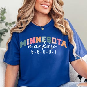 Puede incluir: Una camiseta azul real con el texto "MINNESOTA mankato" en un diseño colorido y arqueado. Debajo del texto está la secuencia numérica "5.6.0.0.1". La camiseta es de un material suave.