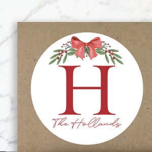 Könnte beinhalten: Runder weißer Aufkleber mit einer roten Schleife und grünen Stechpalmenblättern. Der Buchstabe "H" befindet sich in der Mitte mit dem Text "The Hollands" darunter.