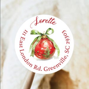 Może przedstawiać: Okrągła biała naklejka z akwarelowym wzorem bombki choinkowej. Czerwona ozdoba ma zieloną kokardę i gałązkę sosny. Naklejka zawiera imię "Soretta" i dane adresowe: III East London Rd., Greenville, SC 29303.
