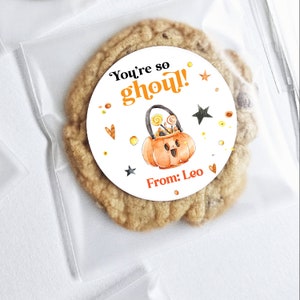 Pode incluir: Um adesivo redondo branco com uma ilustração de abóbora e o texto "You're so ghoul!" e "From: Leo". O adesivo está em um biscoito marrom.