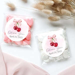 May include: Two clear cellophane bags filled with pastel pink and white candy. Each bag has a round sticker with a watercolour illustration of two red cherries with a pink ribbon. The sticker also includes the text "Olivia Jane" and "April 24, 2025".
