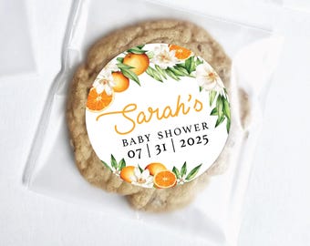 Puede incluir: Una galleta redonda en una bolsa de plástico transparente. La galleta tiene una pegatina blanca con el texto "Sarah's BABY SHOWER 07|31|2025" y un diseño de naranjas, flores blancas y hojas verdes. La galleta es de color marrón claro.